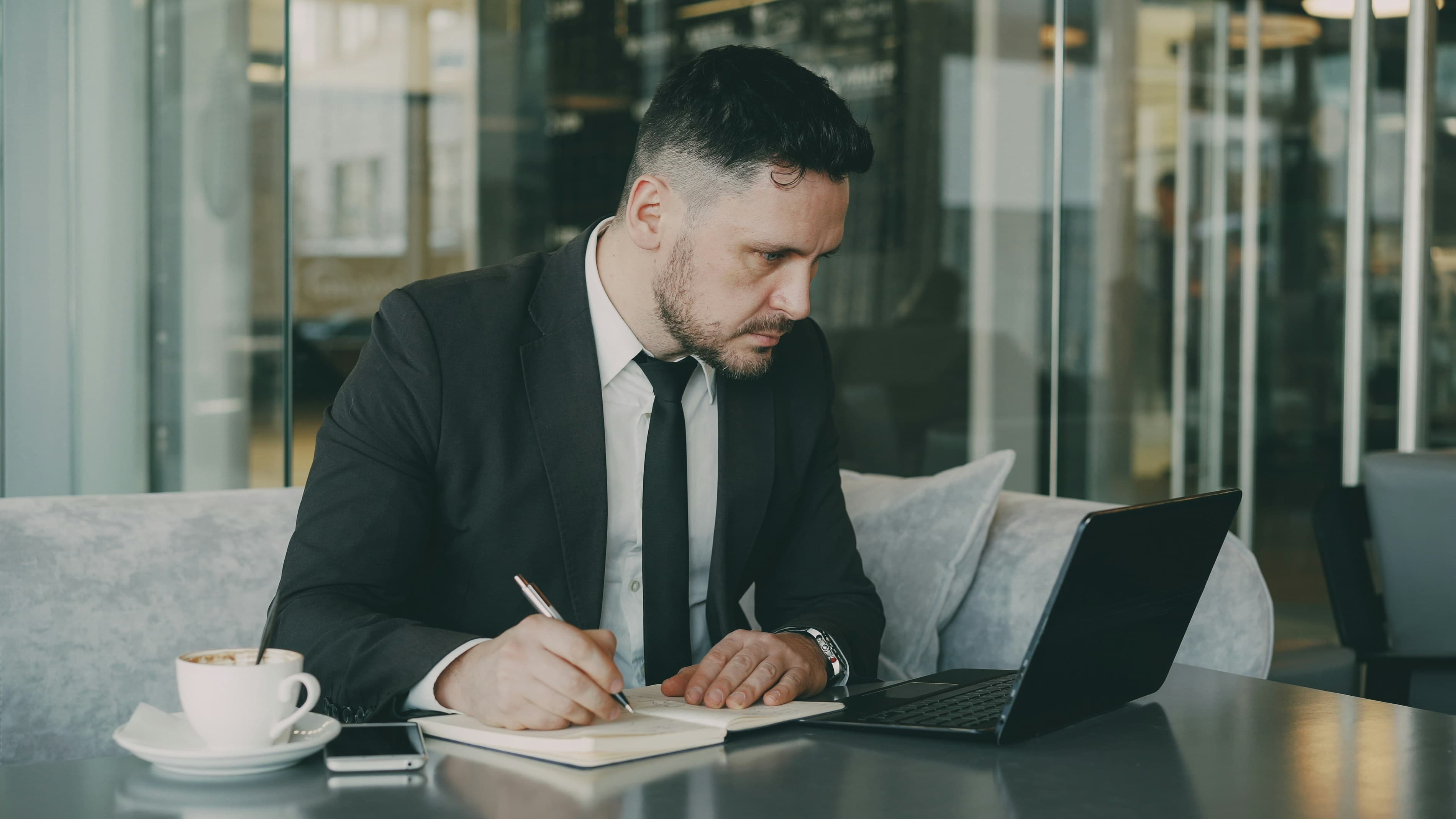 A professional writing in a notebook beside a laptop and coffee